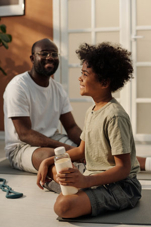 Happy adorable boy with bottle of water enjoying break after workout while sitting on fitness mat and interacting with his fatherの写真素材