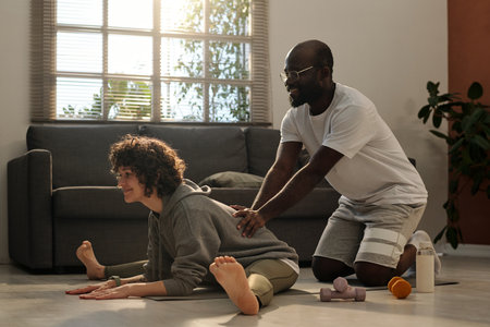 Happy young man in activewear pressing back of his wife doing stretching exercise while standing on sqauts in front of her during workoutの写真素材