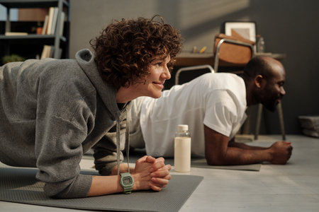 Side view of young brunette woman in grey activewear and her African American husband standing in plank on mats suring workoutの写真素材