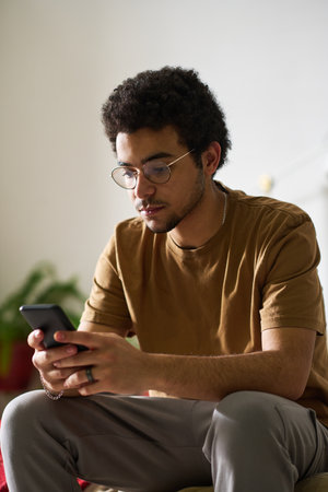 Vertical image of teenage boy in eyeglasses reading message on his smartphone in bedroom