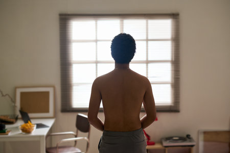 Rear view of young shirtless man in depression sitting in the room and looking at windowの写真素材