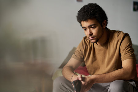 Sad teenage boy waiting for phone call while sitting on bed with smartphone in his hands