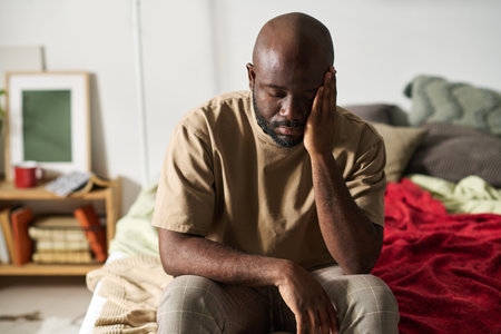 African American man sitting on bed in depression with his eyes closed and having headache