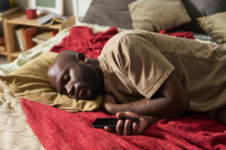 Tired African American man sleeping on bed with smartphone in his hand waiting for callの写真素材