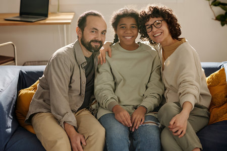 Portrait of happy parents sitting together with their adopted daughter on sofa and smiling at cameraの写真素材