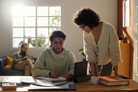 Young mother helping her adopted daughter with her study, she sitting at desk and making notes in...