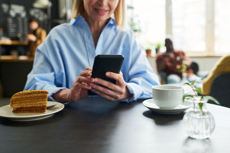 Close-up of mature female consumer using smartphone by table served with cup of tea and piece of...