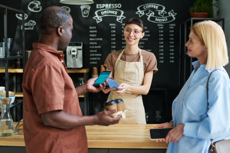 Mature African American man paying for coffee by smartphone in cafe while holding mobile gadget over payment terminal held by clerkの写真素材