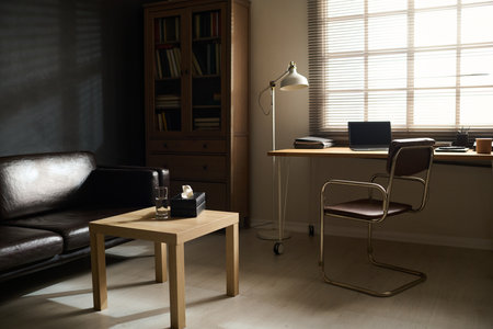 Workplace of psychologist by window and small square wooden table by black comfortable leather couch...