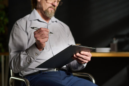 Close-up of mature male psychologist holding clipboard and pen while sitting on chair in front of camera and making notes in documentの写真素材