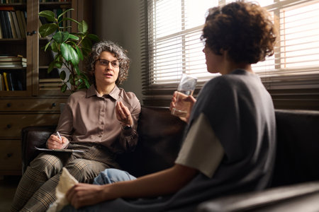 Confident mature psychologist consulting young stressed female patient with glass of water sitting on couch in front of her during sessionの写真素材