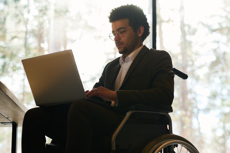 Young multi-ethnic businessman with disability using laptop while sitting in wheelchair in front of camera and preparing new projectの写真素材