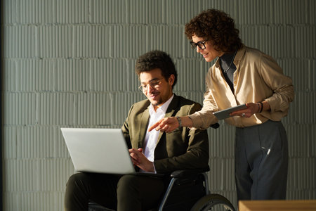 Young businesswoman with tablet bending over male colleague in wheelchair and pointing at screen of laptop on his kneesの写真素材