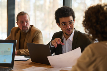 Young confident male economist with financial paper looking at female colleague during their conversation at meeting in boardroomの写真素材