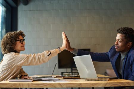Successful young male and female colleagues giving each other high-five over workplace with laptops,...