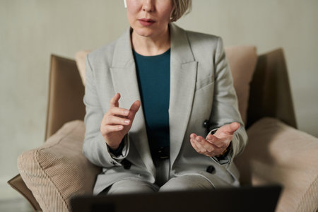 Close-up of confident psychologist in grey suit sitting in armchair in front of laptop during online...