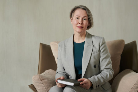 Successful mature psychologist wearing grey elegant suit sitting in armchair against grey wall in...
