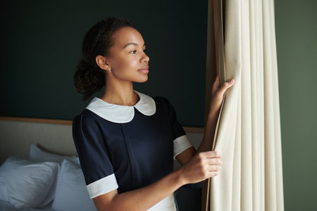 Young pretty housemaid in dark blue dress opening curtains in hotel room in the morning while...