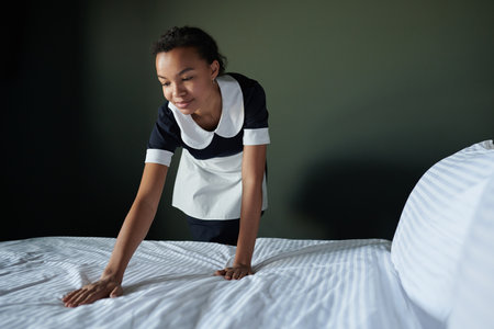 Young African American chambermaid bending over comfortable double bed while changing blanket and...