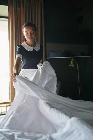Young chambermaid in uniform standing by double bed in hotel room and changing blanket and other...