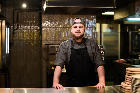 Young confident owner of pizzeria standing by large table in the kitchen of his restaurant and...