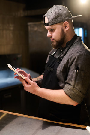 Side view of young serious cook in uniform scrolling through online recipes on tablet screen while...