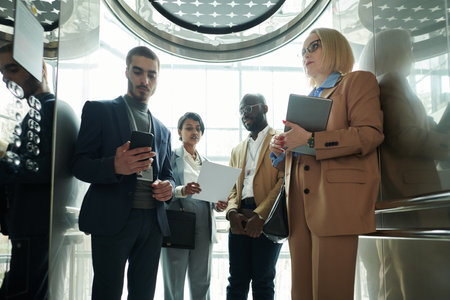 Group of intercultural entrepreneurs revising points of their reports in smartphone and on paper while standing in moving elevatorの写真素材