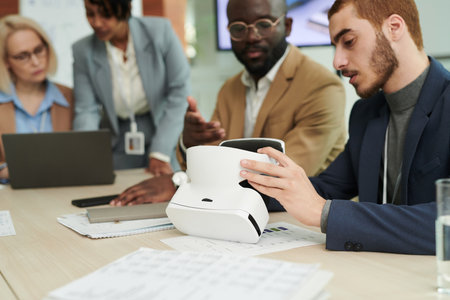 Focus on young businessman making presentation of vr headset to African American male colleague while sitting against two businesswomenの写真素材