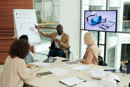 Group of businesswomen listening to male speaker in wheelchair pointing at whiteboard during explanation of graphic data at seminarの写真素材