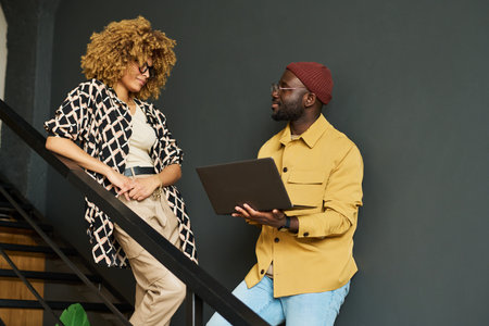 Two young African American colleagues in casualwear standing on staircase and discussing online data...