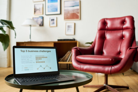 Laptop with financial graphic data on screen standing on small round table in front of camera against armchair and pictures hanging on wallの写真素材