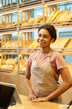 Young smiling clerk of bakery shop in apron looking at camera while standing by counter desk with computer against display with breadの写真素材