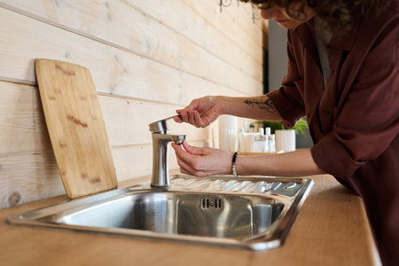 Hands of young housewife in brown shirt trying to open metallic faucet over sink while standng by wooden counter in the kitchen at homeの写真素材