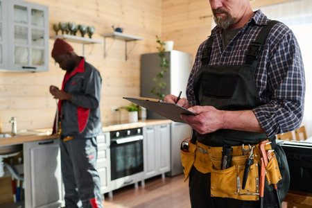 Cropped shot of mature male worker of repair service company making notes in document against his colleague standing by kitchen counterの写真素材