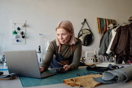 Blond mature owner of leatherworking shop looking at laptop screen while standing by workplace and...