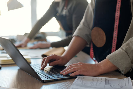 Close-up of young craftswoman pressing keys of laptop keyboard while standing by workplace and checking new online orders against colleagueの写真素材