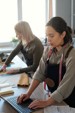 Focus on young brunette female leatherworker typing on laptop keyboard while checking online orders of clients against mature colleagueの写真素材