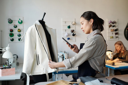 Side view of young female fashion designer scrolling in smartphone or taking photo of new leather...
