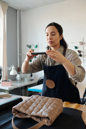 Young craftswoman in apron taking photo of new handmade leather handbag on smartphone while standing by workplace in studioの写真素材