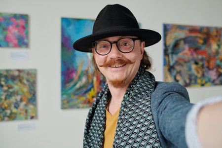 Artist in hat smiling at camera while making selfie portrait against his paintings in galleryの写真素材