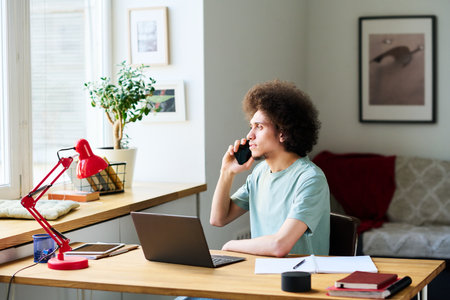 Young man with curly hair sitting at his workplace with laptop and having conversation on mobile...