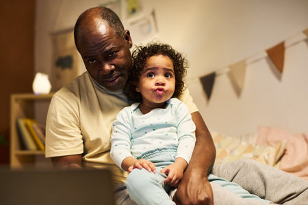 African American dad using laptop while playing with his child in bedroom