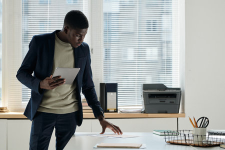 Young serious businessman with tablet standing by desk with financial documents in openspace office and looking through papersの写真素材
