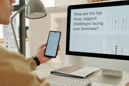 Focus on computer monitor with data on desk and smartphone with blank screen in hand of young male...