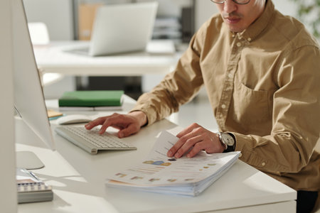 Cropped shot of young businessman in beige shirt working with financial documents and using computer...
