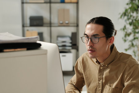 Young serious analyst in eyeglasses and beige shirt sitting by workplace in front of computer and...