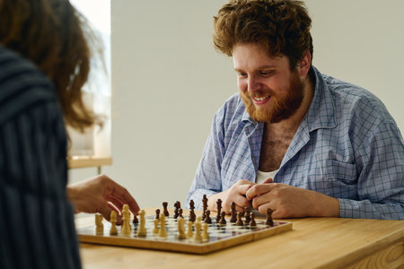 Young smiling male patient of mental clinics looking at chessboard while game partner making move...
