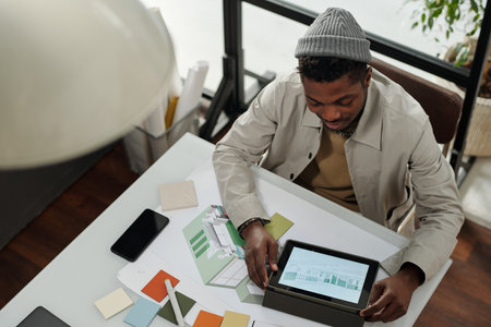 Above view of young African American specialist using tablet with graphic sketches on screen while sitting by desk and working over new projectの写真素材