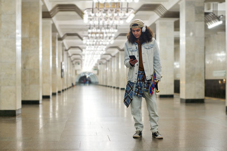 Teenage guy in denim casualwear texting in mobile phone while standing against long subway tunnel...