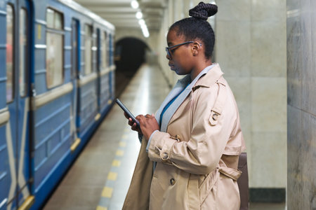 Side view of young black woman scrolling in smartphone or watching video while standing in front of moving blue train at subway stationの写真素材